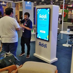 Promotor da Addi conversando com cliente no stand, ao lado de Totem, com aplicativo e adesivo da Addi