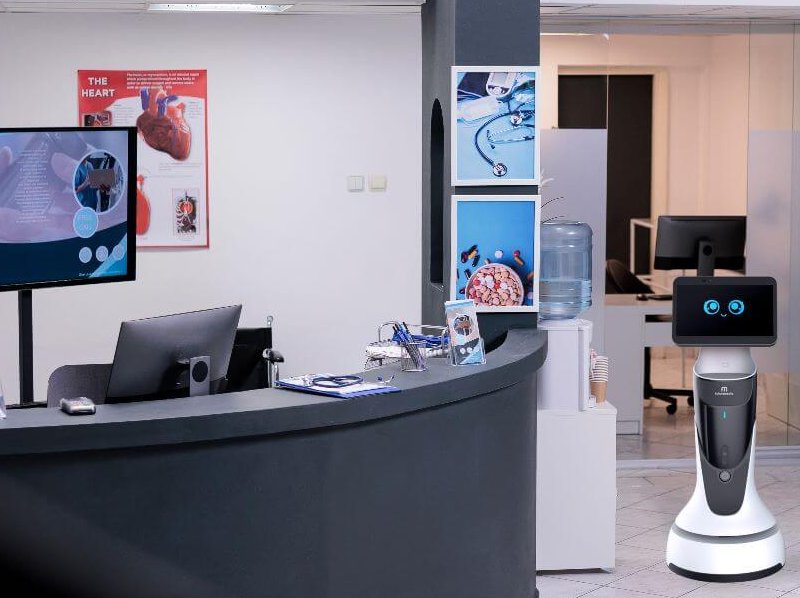 Robô Buddy com carinha sorridente em sua tela, ao lado de recepção de hospital